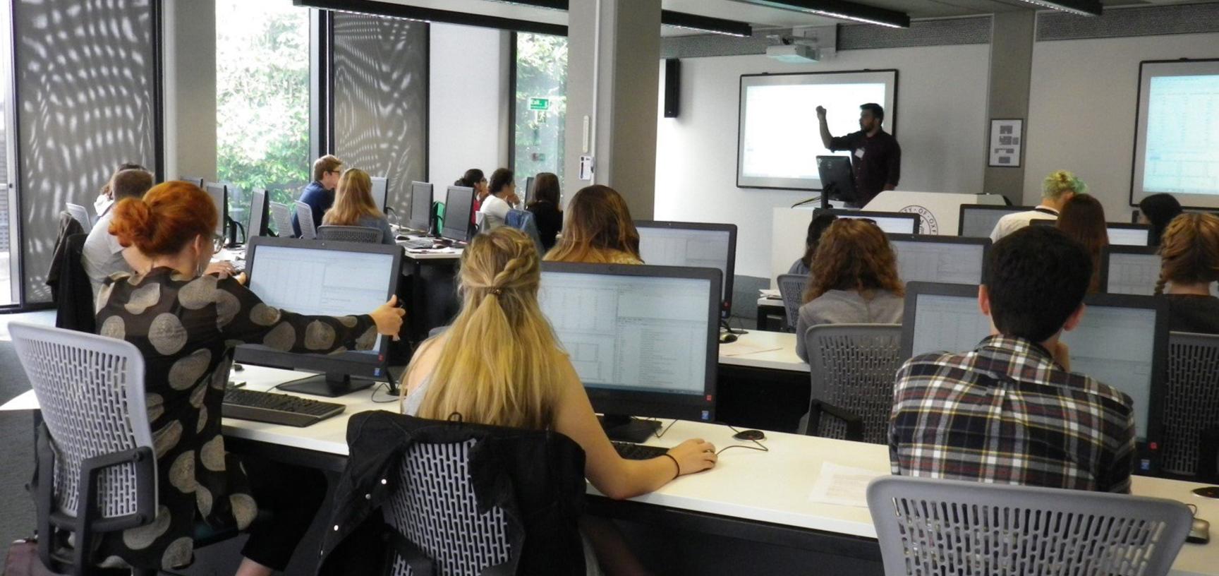 View from the back of computer lab with students in front of desktops and lecturer at the front next to projector screen