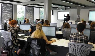 View from the back of computer lab with students in front of desktops and lecturer at the front next to projector screen