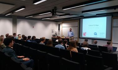 Wide view of lecture hall with lecturer standing next to large screen in front of Summer Institute students