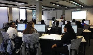 View from the back of computer lab with students in front of desktops and lecturer at the front next to projector screen