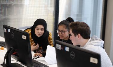 Two female students and one male student sitting in front of desktop computer and discussing while looking at the screen